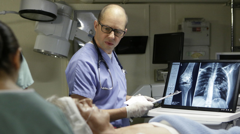 Jon Cryer as Dr. Taft in blue scrubs standing above Mark Harmon as Gibbs in hospital bed in NCIS