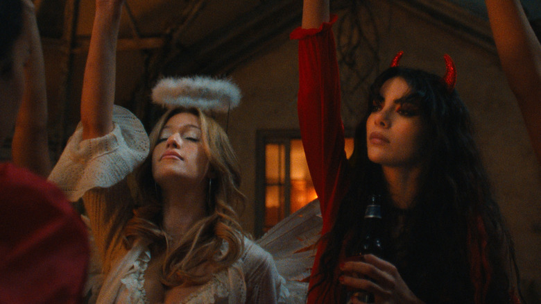 In a dimly lit house, a blonde woman is dressed in a white lace top with feathery wings and a fluffy halo headband with her eyes closed, and a woman with dark hair wearing a red top and red devil horns is holding a bottle of beer.