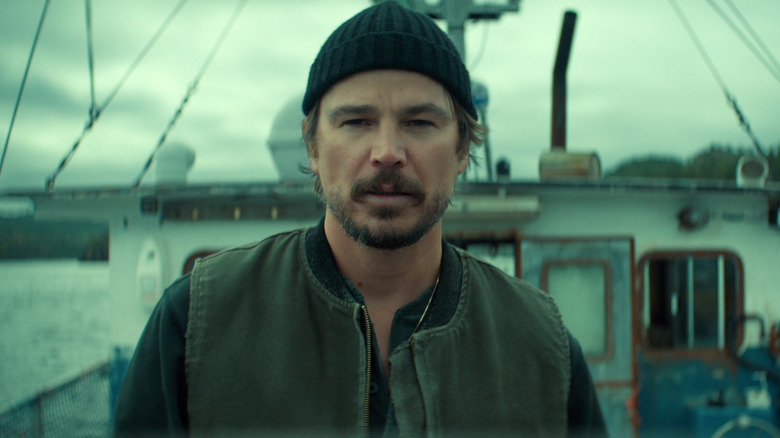 A man with a graying brown beard wearing a black beanie hat, a zipped-up, sleeveless gray vest, and a black sweater underneath stands on the deck of a large fishing boat.