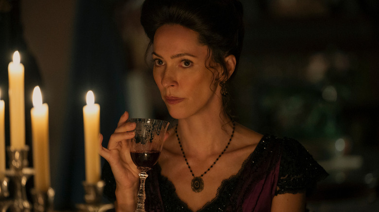 A woman in period costume with her hair tied in an up-do is holding a glass of red wine, and there are candles in the foreground.