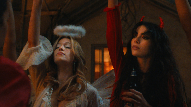 In a dimly lit house, a blonde woman is dressed in a white lace top with feathery wings and a fluffy halo headband with her eyes closed, and a woman with dark hair wearing a red top and red devil horns is holding a bottle of beer.