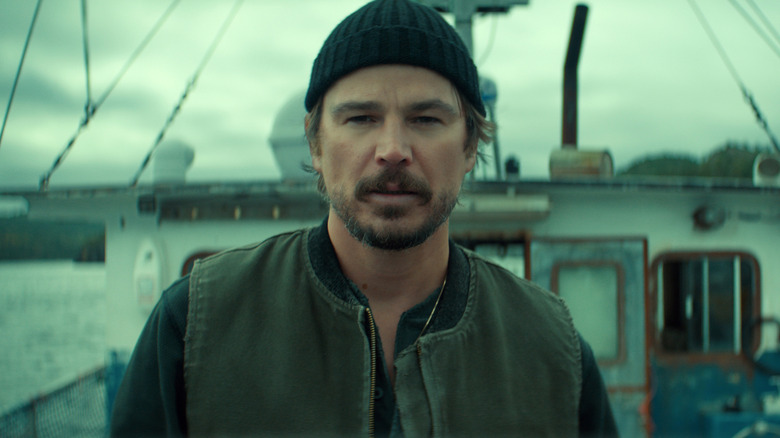 A man with a graying brown beard wearing a black beanie hat, a zipped-up, sleeveless gray vest, and a black sweater underneath stands on the deck of a large fishing boat.