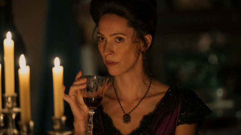 A woman in period costume with her hair tied in an up-do is holding a glass of red wine, and there are candles in the foreground.
