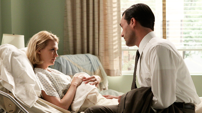 Don Draper sits beside a tired-looking Betty Draper, propped up, holding her baby, with beige curtains in the background