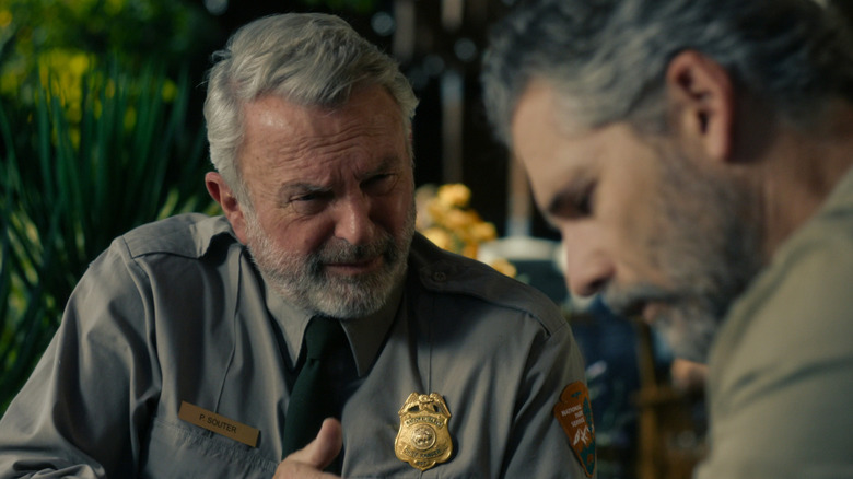 On the left is Sam Neill with gray hair and a beard wearing a Park Ranger Chief uniform, on the right is Eric Bana with graying black hair wearing a khaki shirt — Bana is out of focus.