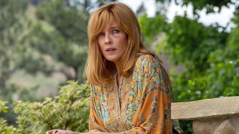 Beth Dutton rolünde Kelly Reilly, siyah gözlü, sarı ve turuncu çoklu desenli bir elbise giyiyor, Yellowstone'da dışarıya bakan bir bankta oturuyor