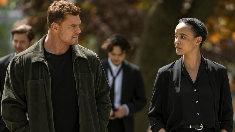 A man and a woman are walking through a park. On the right the man has short brown hair and is wearing a green over shirt with a black t-shirt. On the right the Black woman has her hair tied up and is wearing a black blazer over the top of a black button-up shirt.