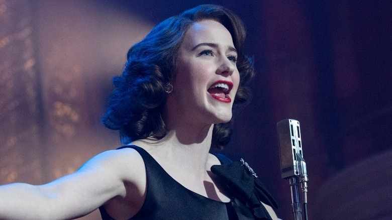 Midge Maisel standing on stage at a microphone
