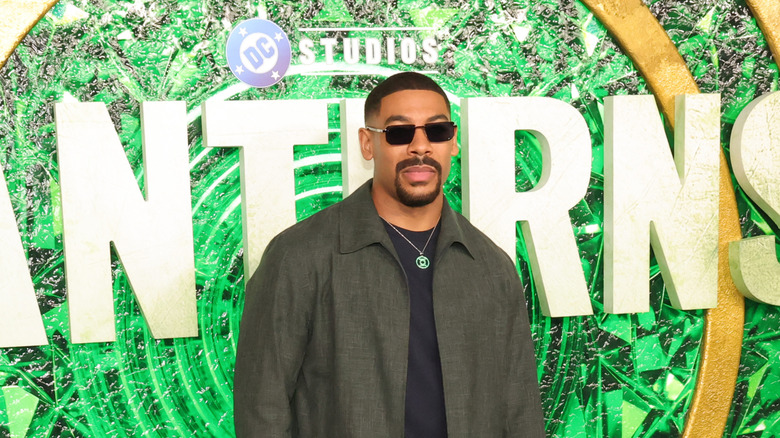 A Black man with short black hair and a goatee is wearing  dark sunglasses and a dark green overshirt with a black t-shirt underneath. There is a silver chain around his neck with a Green Lantern symbol attached. Behind him is the DC Studios logo on top of a large "LANTERNS" logo in white  with a shattered green crystal design in the background..