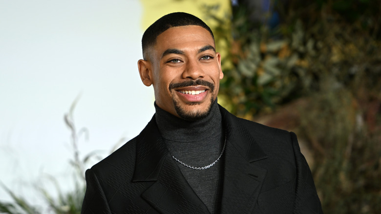 A Black man with short black hair and a goatee is wearing a black suit with a dark gray turtle neck sweater and a small silver chain.
