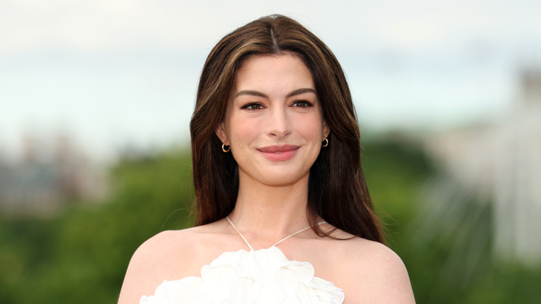 A woman with long brown hair smiling at something off-camera. She is wearing two small gold earrings, as well as a frilly white dress that has a small white strap looped around her neck.