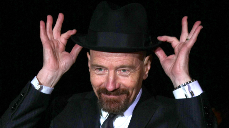 Bryan Cranston keçi sakallı, siyah bir takım elbise giyiyor ve siyah bir fötr şapkayla kravat takıyor. Sanki şapkayı yeniden kafasına yerleştiriyormuş gibi ellerini şapkanın üzerinde tutuyor.