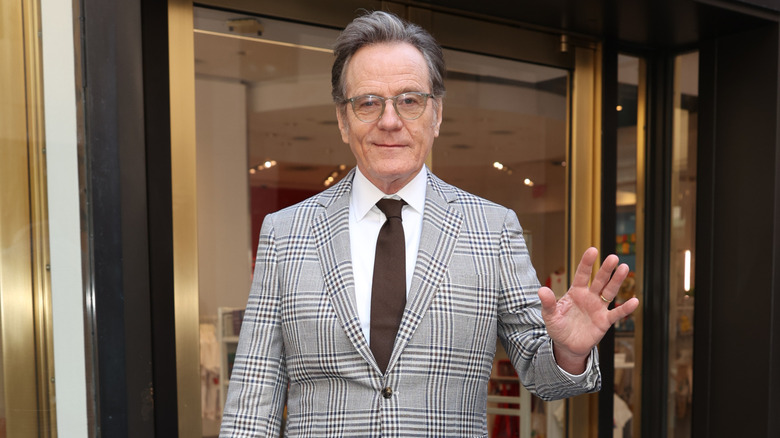 Bryan Cranston, gri saçlı, gümüş gözlüklü, gri kareli takım elbise ceketli, beyaz gömlekli ve kahverengi kravatlı.