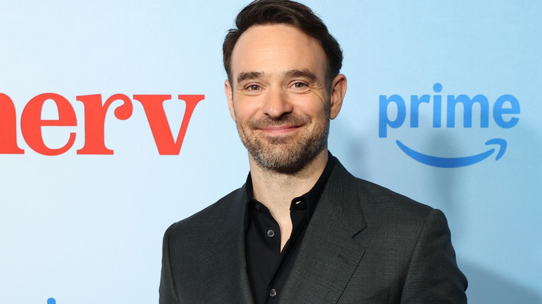 A man with short brown hair smiling for the camera, he has short brown stubble. He's wearing a dark gray suit and a black shirt. He's stood against a blue background with a red logo for "Merv" and a blue logo for "Prime."
