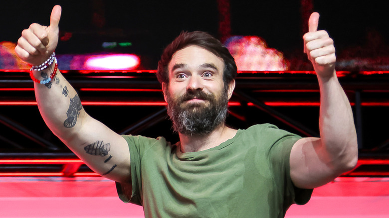 A man with messy brown hair and a large brown graying beard on stage. he's holding both his hands up with his thumbs up. He's wearing a green t-shirt. There are multiple friendship bracelets around his right arm, and he has three black tattoos on the same arm.