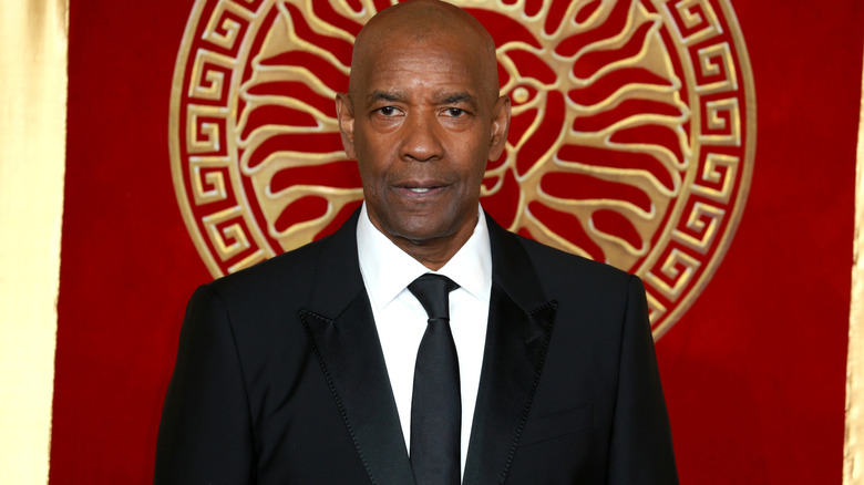 Denzel Washington wearing a black suit and tie. He's clean shaven and has a bald head. There is a red and gold pattern on the wall behind him.