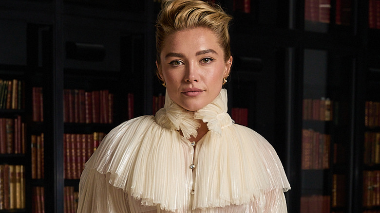 Flornece Pugh with blond hair coffed up. She's wearing a white frilly blouse and she's stood in a library.