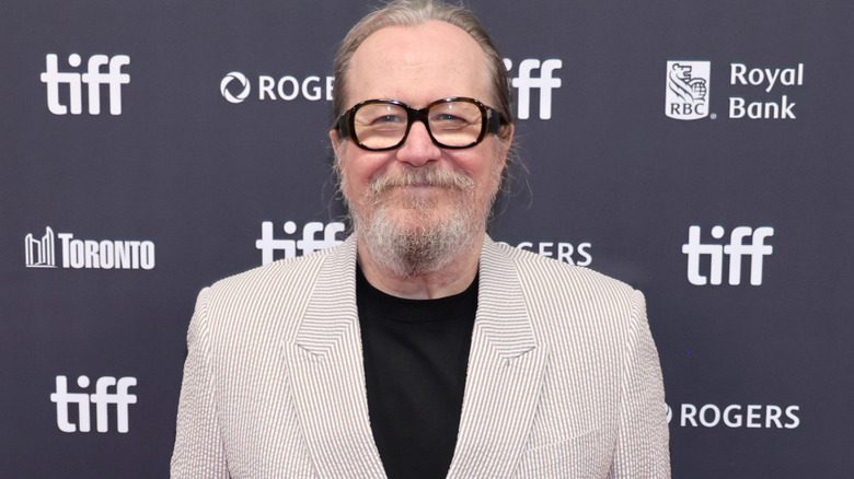 Gary Oldman with long gray hair tied behind his head, he's also got a gray beard and is wearing brown glasses. He's wearing a gray striped blazer with a black t-shirt. He's stood against a black background with the Toronto International Film Festival logo on it.