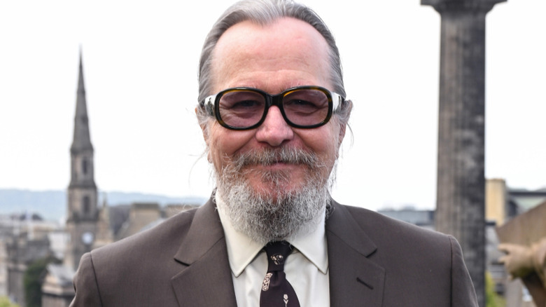 Gary Oldman with long gray hair tied behind his head, he's also got a gray beard and is wearing brown glasses. He's wearing a brown suit with a white shirt and a brown tie.