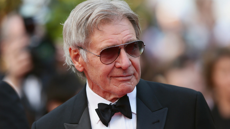 Harrison Ford with gray hair wearing a black tuxedo and aviator sunglasses.