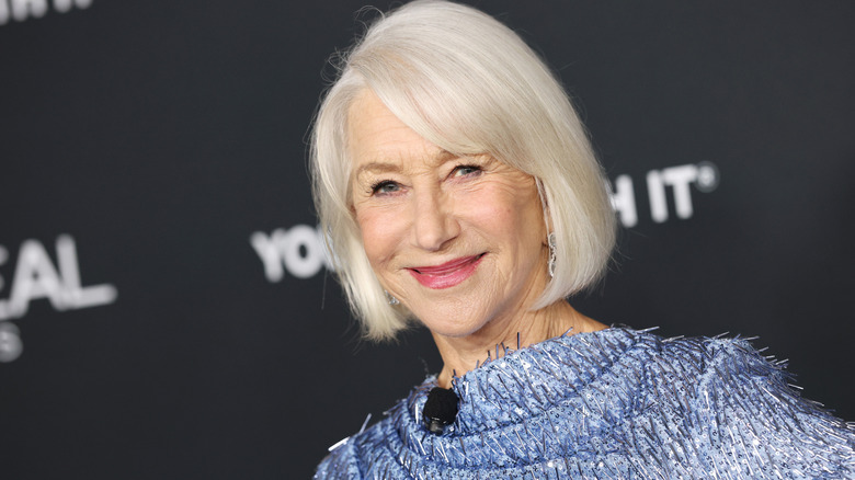 Helen Mirren with white hair ina. bob wearing a blue glittery shawl, she's stood against a black background.