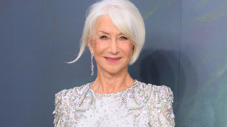 Helen Mirren with white hair wearing diamond earrings and a diamond encrusted dress. She's stood against a green-blue background.