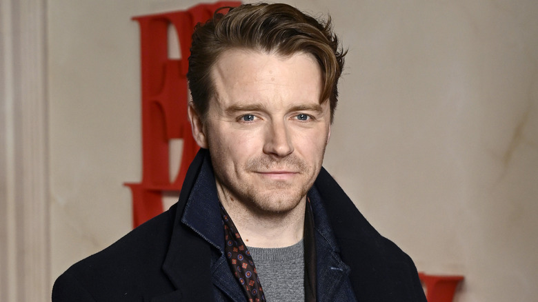 A man with coiffed brown hair with a curled fringe looks at the camera. He's got short stubble and is wearing a navy coat with the collar up with a denim under shirt and a gray sweater. There is a marble background behind him with a bright red logo on it.