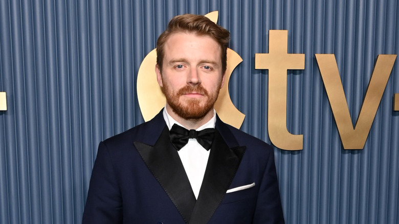 A man with slicked back ginger hair is wearing a navy double breasted tuxedo with a black tie and a white shirt. There is a blue lined background behind him with a gold Apple TV logo on it.