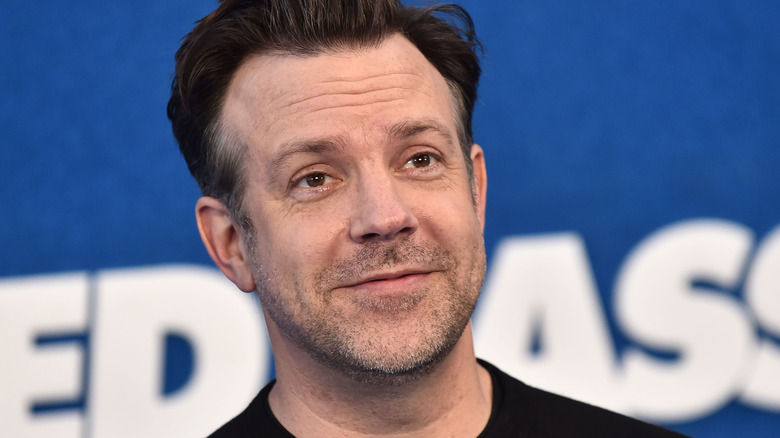 Jason Sudeikis with swept-back brown hair and short stubble. He's wearing a black top and he's stood against a blue backdrop with a white "Ted Lasso" logo on it.