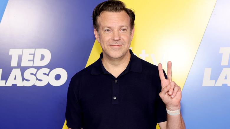Jason Sudeikis with swept-back brown hair. He's wearing a black polo top against a blue and yellow backdrop. He's holding up a peace sign.