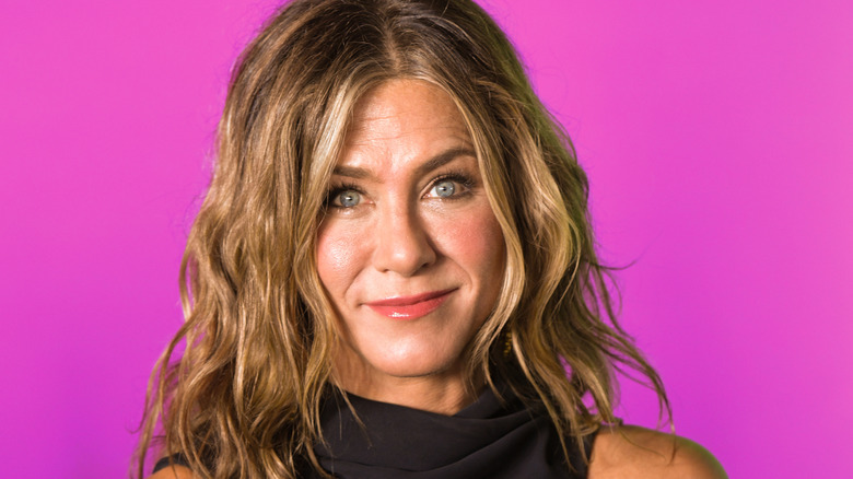 Jennifer Aniston with curly brown/blond hair while wearing a purple dress. She's stood against a bright pink backdrop.