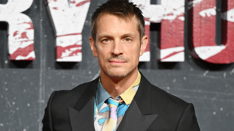 Joel Kinnaman with short brown hair and a short goatee. He's wearing a black suit with a yellow, blue, purple, and gray patterned shirt & tie.