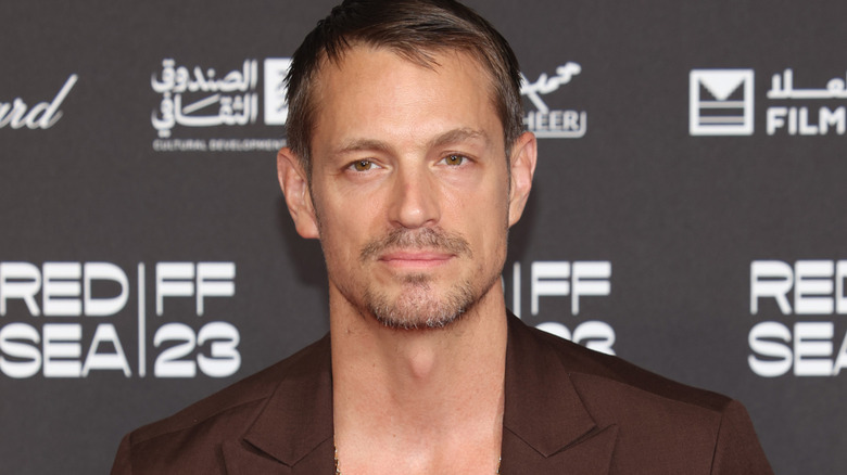 Joel Kinnaman with short brown hair and goatee wearing a brown suit against a black background with the white logo for the Red Sea Film Festival 2023 on it.