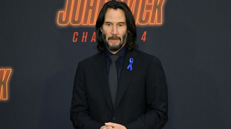 Keanu Reeves with long dark hair and a graying beard. He's wearing a black suit and tie with a navy blue shirt. A blue ribbon is pinned to his lapel. The orange logo for "John Wick Chapter 4" is on a black wall behind him.