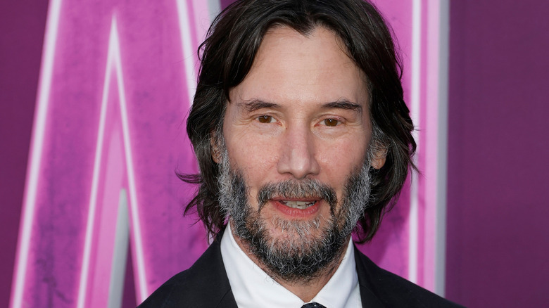 Keanu Reeves with long dark hair and a graying beard. He's wearing a black suit with a white shirt. He's stood against a neon pink wall.