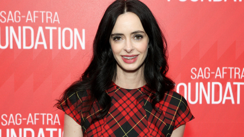 Krysten Ritter with long black hair wearing a red and black tartan dress, she's stood against a red background with the SAG-AFTRA Foundation logo in white text behind her.
