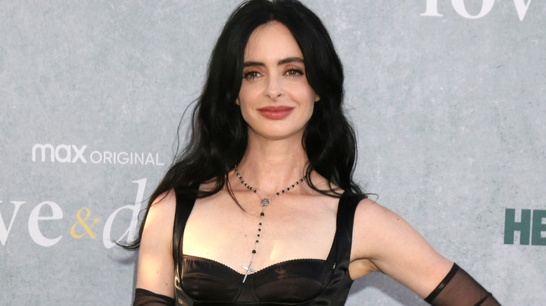 Krysten Ritter with long black hair wearing a black leather-look dress. There's black-beaded crucifix around her neck, and she's stood against a gray background.