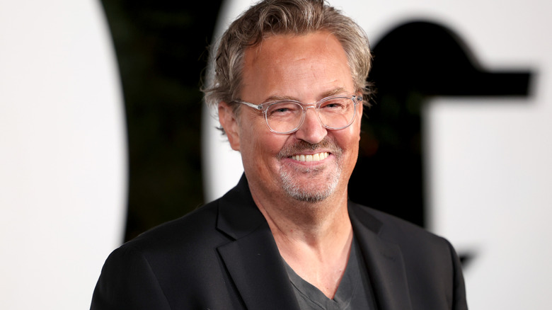 Matthew Perry with gray hair and a gray goatee wearing clear frame glasses and a black blazer.
