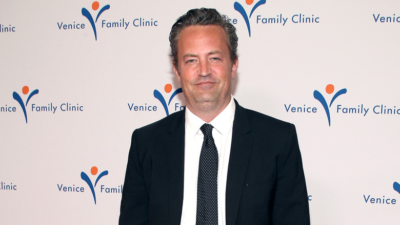 Matthew Perry with graying hair wearing a dark suit and a dotted tie with a white shirt.