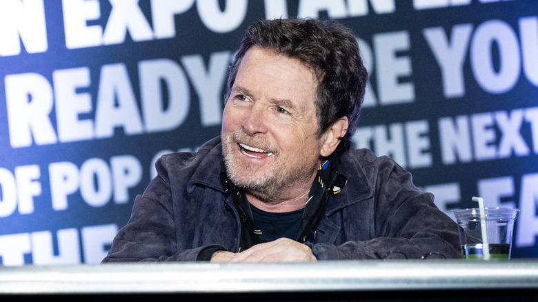 Michael J. Fox with brown hair and a graying a beard. He's wearing a blue suede jacket and he's sat at a table. There is a wall behind him with large white text.
