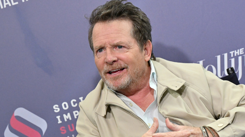 Michael J. Fox with brown hair and a graying a beard. He's wearing a beige jacket with a white button-up shirt. There is a blue wall behind him.