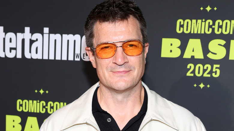 Nathan Fillion with short dark hair wearing a white jacket, a black shirt and orange sunglasses. He's stood against a black wall featuring a white Entertainment Weekly logo and a yellow Comic-Con logl.