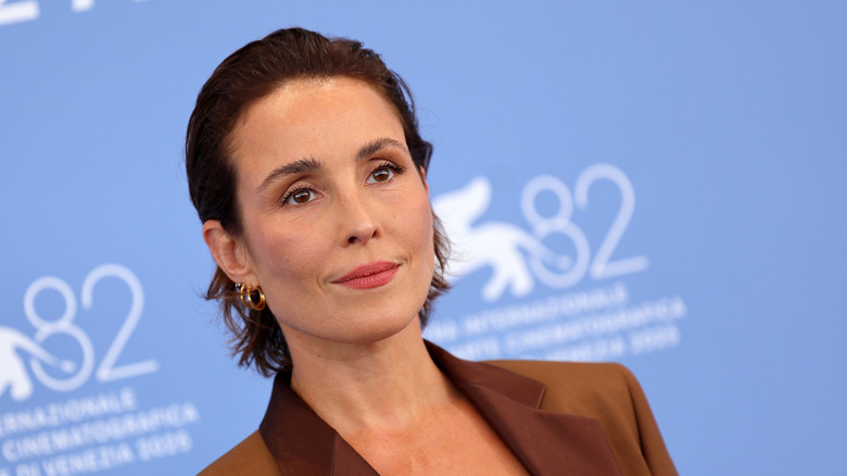 Noomi Rapace at a press event smiling and wearing a brown blazer