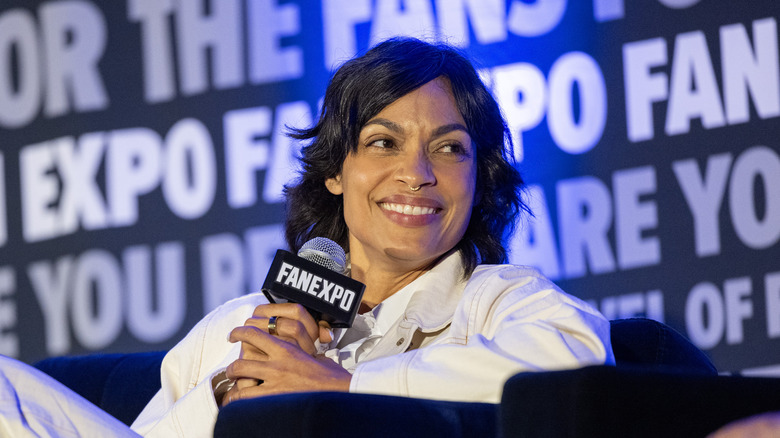 A Black woman with shoulder-length black hair is sitting on a couch holding a microphone with a white "FANEXPO" logo on it. She's wearing a white denim jacket and is wearing a small septum ring in her nose. There is a wall of text behind her featuring the "FAN EXPO" logo.