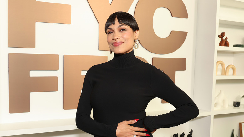 A Black woman with short cropped black hair wearing a long-sleeve black turtleneck. She has red nails and is wearing gold earrings with a small gold septum ring in her nose. There is large gold text on the white wall behind her reading "FYC FEST."