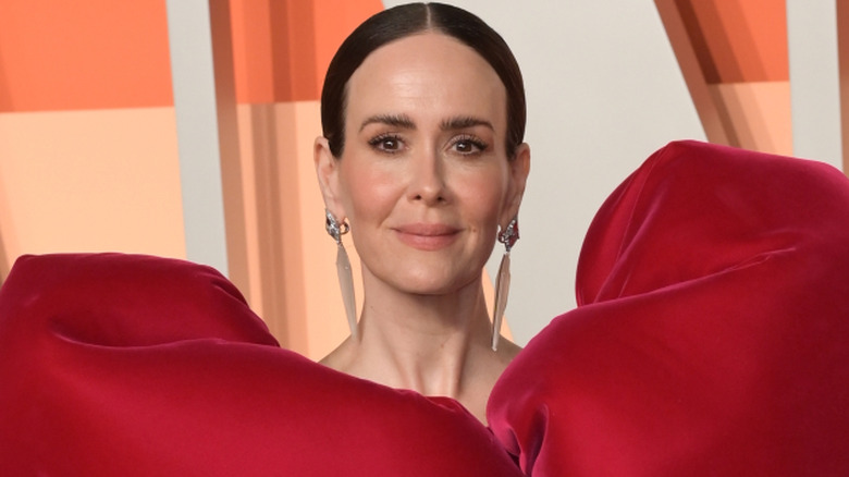 Sarah Paulson with tied-back brown hair. She's wearing long earrings and a large puffy red dress.