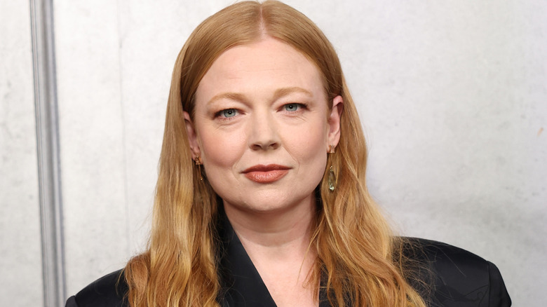 Sarah Snook with long ginger hair, she's wearing a black dress and is stood against a stony background.