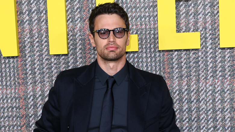 A man with neat brown hair and short stubble is wearing black sunglasses with a navy blue coat, shirt and tie. He's stood against a checked, fabric background made of white, green, and light red., There is also large yellow letters above him that are cropped by the image.