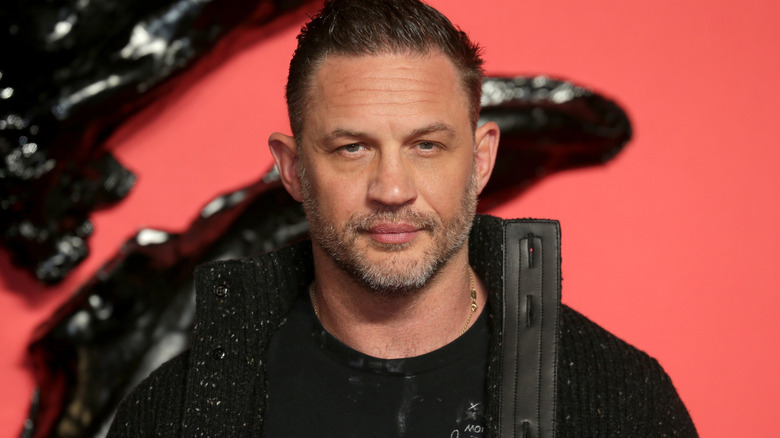 Tom Hardy with slicked back brown hair and a short beard wearing a black cardigan with a large leather collar. He's wearing a black t-shirt underneath and he's stood against a red and black background.