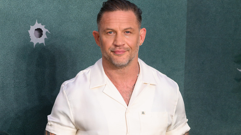 Tom Hardy with slicked-back brown hair wearing a white cuban-collared shirt. He's stood against a dark greeny/blue background with a bullet hole design above his right shoulder.